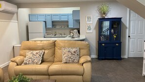 A living room with a beige sofa, a coffee table with a plant, a kitchen area with blue cabinets, and a blue cabinet with glass doors. - Adorable 1-bedroom barn in Mineola, East Texas also known as Lake Country (Mineola)