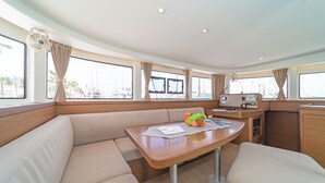 Interior d'un vaixell amb zona de menjador, seients amb coixins, taula amb un plat de fruita i quadre de comandament.