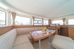A boat interior with a dining area, cushioned seating, a table with a fruit bowl, and a control panel. - Sun-Kissed Sailing Adventure in the Dodecanese Islands (Kos)