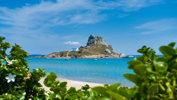 A coastal landscape with a prominent rock formation, clear blue water, and a small building on a hill. - Sun-Kissed Sailing Adventure in the Dodecanese Islands (Kos)