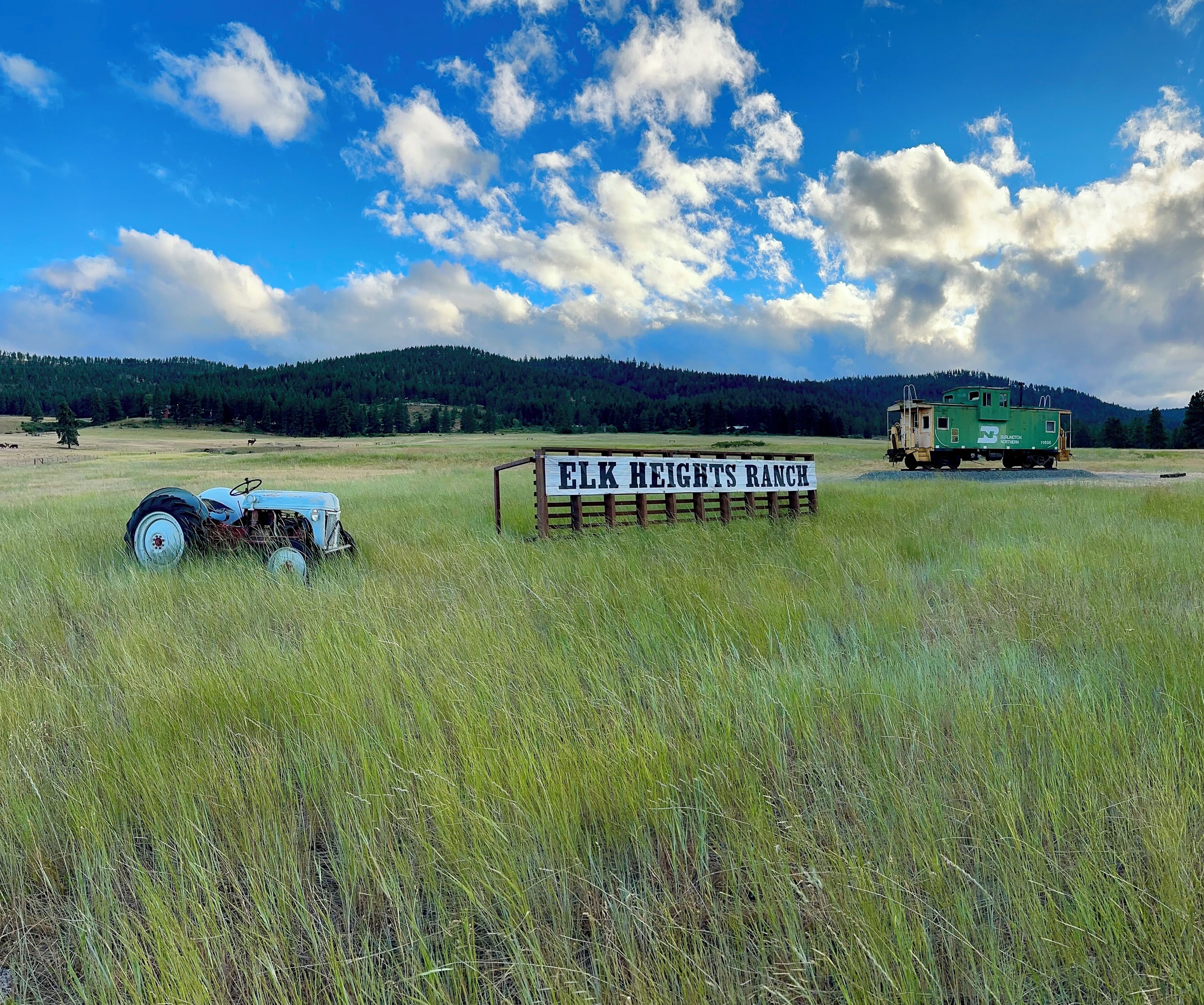 Ein Schild mit der Aufschrift 'ELK HEIGHTS RANCH' in einem grasbewachsen Feld, im Hintergrund ein blauer Traktor und ein grüner Zugwagen.