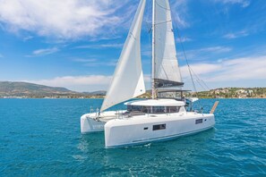 A white catamaran with a cabin and a sail, floating on a calm sea with a distant shoreline.
