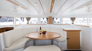 A boat interior with a round table, cushioned seating, and windows with curtains. - Sun-Kissed Sailing Adventure in Islas Sarónicas (Alimos)