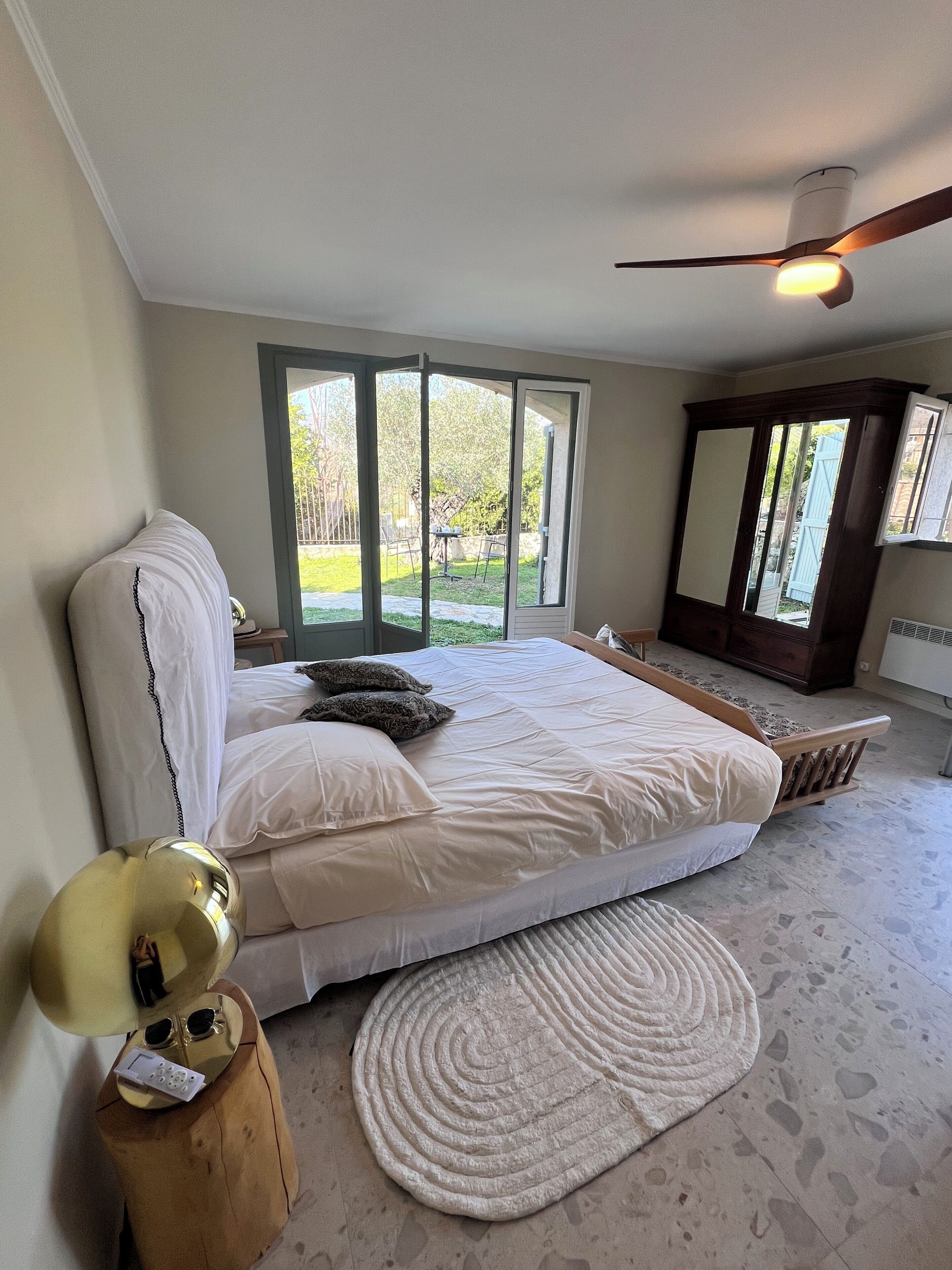 A bedroom with a bed, a bedside table, a mirror, and a view of the outdoors.