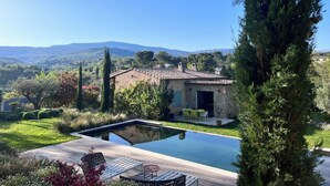 Una piscina con una casa de piedra al fondo, rodeada de vegetación y colinas.