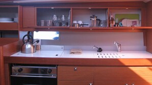 A compact kitchenette with a stove, kettle, sink, and shelves above for storage. - Sun-Kissed Sailing Adventure in Islas Sarónicas (Alimos)