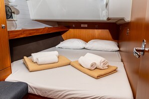 Ein kleines Zimmer mit einem Bett, zwei Kissen, einem Nachttisch, einem kleinen Stuhl und einem fest installierten Ventilator.
