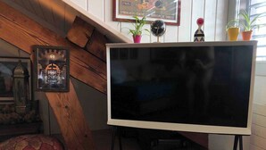 A modern living room with a large flat-screen TV on a stand, wooden beams, a red pouf, a framed picture, and potted plants. - Stylish Loft - Amazing Views (Geneva)