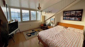 A bedroom with a sloped ceiling, a bed, a desk, a chair, a TV, and a window with blinds. - Stylish Loft - Amazing Views (Geneva)