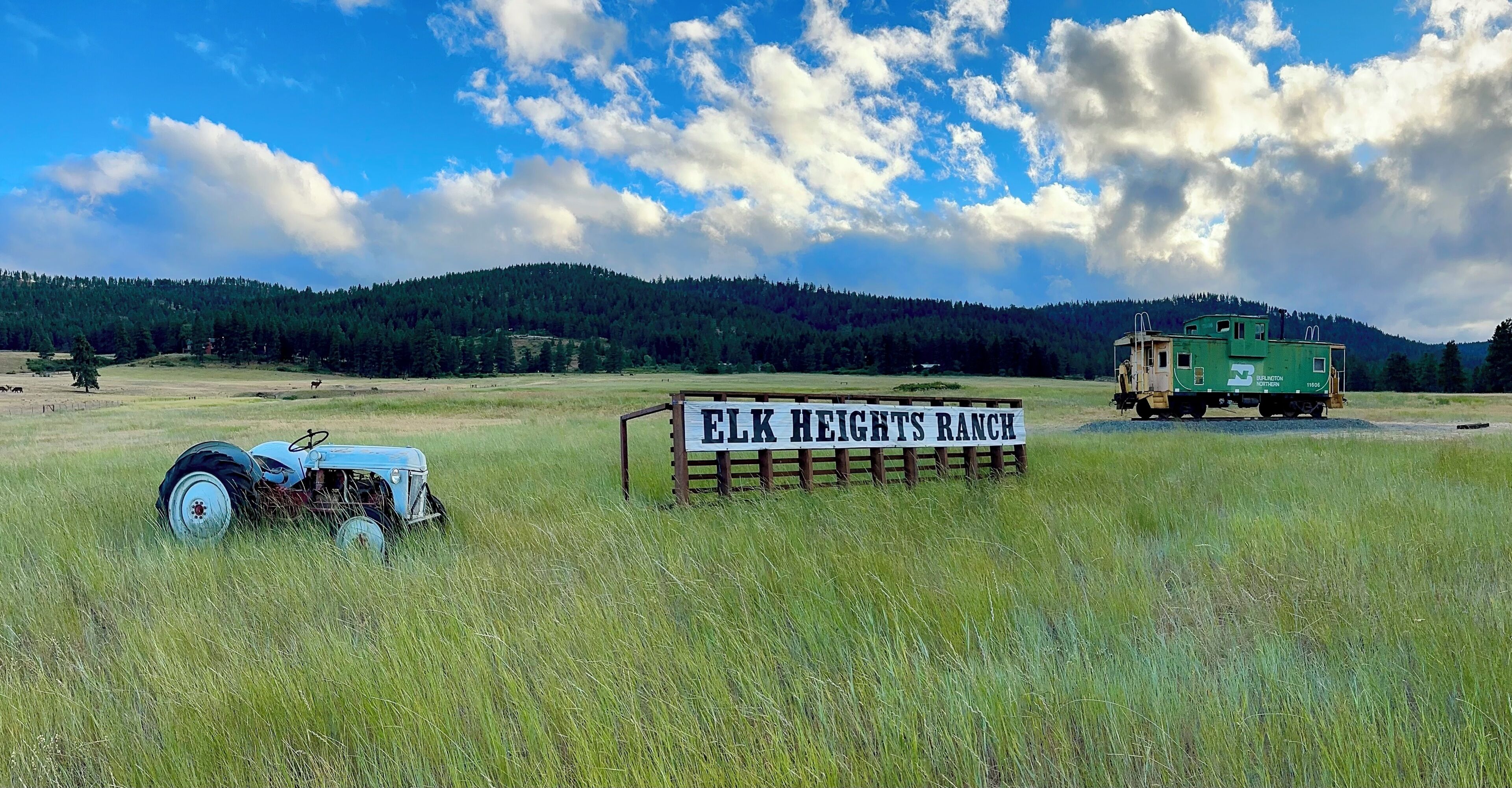 Ländliche Landschaft mit einem Schild mit dem Schriftzug 'ELK HEIGHTS RANCH', einem Traktor und einem grünen Fahrzeug auf dem Feld.