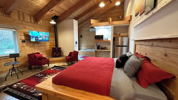 Interior de una cabaña de madera con una cama, sillas, un televisor, chimenea y una zona de cocina.
