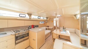 A compact kitchen with a stove, oven, and sink, a small dining area with a table and bench, and a window providing natural light. - Sun-Kissed Sailing Adventure in Islas Sarónicas (Alimos)