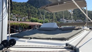 Convés de barco com área de dormir coberta, cordas e vista para uma cidade costeira e colinas.