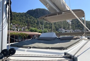 Ein Bootsdeck mit einer überdachten Liegefläche, Seilen und Blick auf eine Küstenstadt und Hügel.