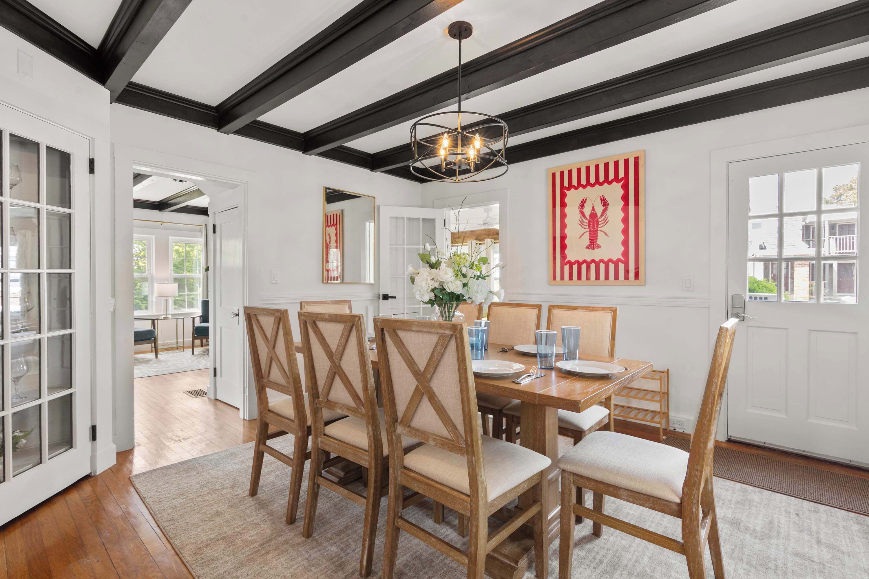 A dining room with a wooden table, chairs, and a chandelier. There is a door leading to another room and a window with a view.