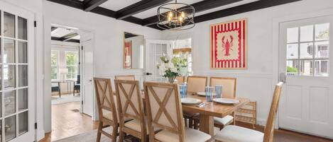 A dining room with a wooden table, chairs, and a chandelier. There is a door leading to another room and a window with a view.