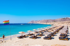 A beach with rows of sun loungers and umbrellas, a flag, and a clear blue sea.