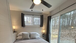 A bedroom with a bed, a ceiling fan, two bedside lamps, a window with curtains, and a view of trees. - Cozy Lofted Cabin #2 at Cataract Lake Getaway (Poland)