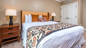 A neatly made bed with a patterned bedspread, two bedside lamps, a wooden nightstand with drawers, and a white door in the background. - Honua Kai 3 Free Cars KBM Resorts Front Alii Suite 3 Units, 6 Bedrooms HK Ml-3146 (Kaanapali)