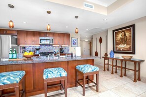A kitchen with wooden cabinets, a granite countertop, a microwave, and a stove. There are three wicker bar stools with blue cushions. - Honua Kai 3 Free Cars KBM Resorts Front Alii Suite 3 Units, 6 Bedrooms HK Ml-3293 (Kaanapali)