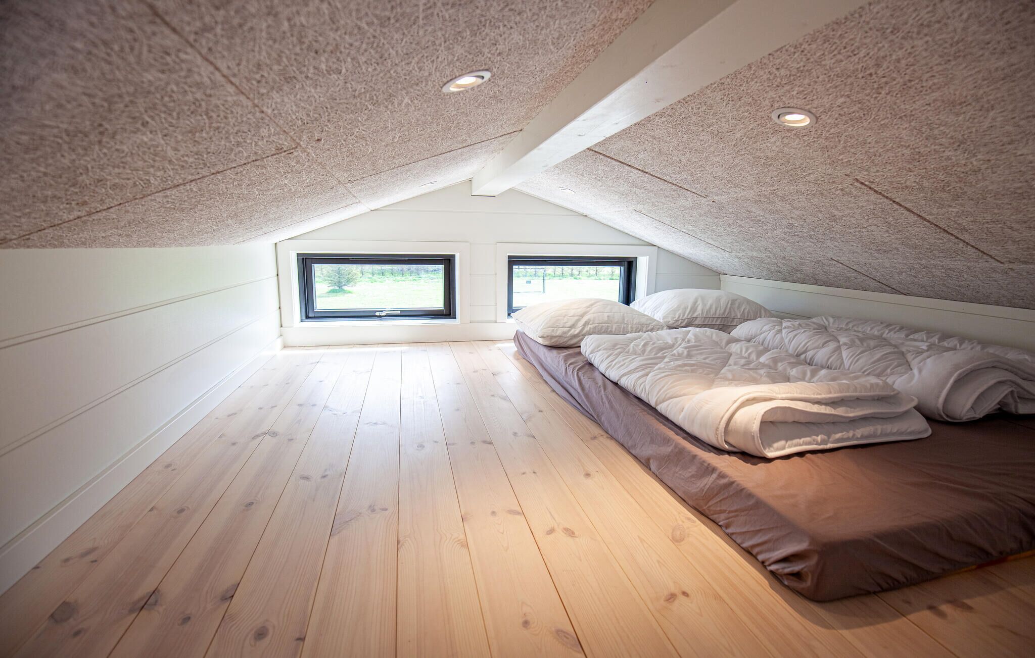 Un dormitorio con techo inclinado, suelo de madera, una cama con ropa blanca y dos ventanas.