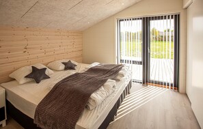 A bedroom with a wooden headboard, a bed with white linens, a brown blanket, and a view of a green landscape through a sliding glass door. - Pet friendly home in Sæby with sauna (Sæby)
