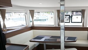 A compact interior with a dining table, cushioned seating, a small refrigerator, and a window with a view of the marina. - Sun-Kissed Sailing Adventure in the Saronic Islands (Laurium)