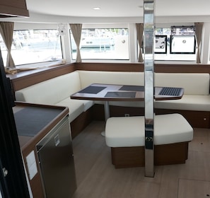A compact interior with a dining table, cushioned seating, a small refrigerator, and a window with a view of the marina. - Sun-Kissed Sailing Adventure in the Saronic Islands (Laurium)