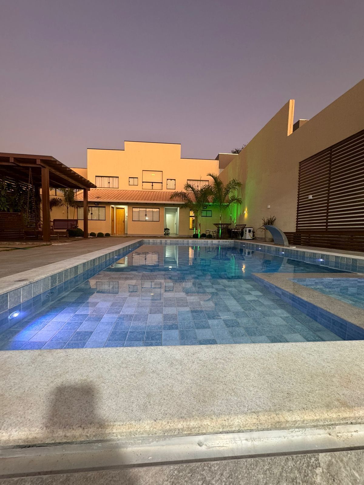 A swimming pool with a tiled deck, a covered seating area, and a building with multiple windows in the background.