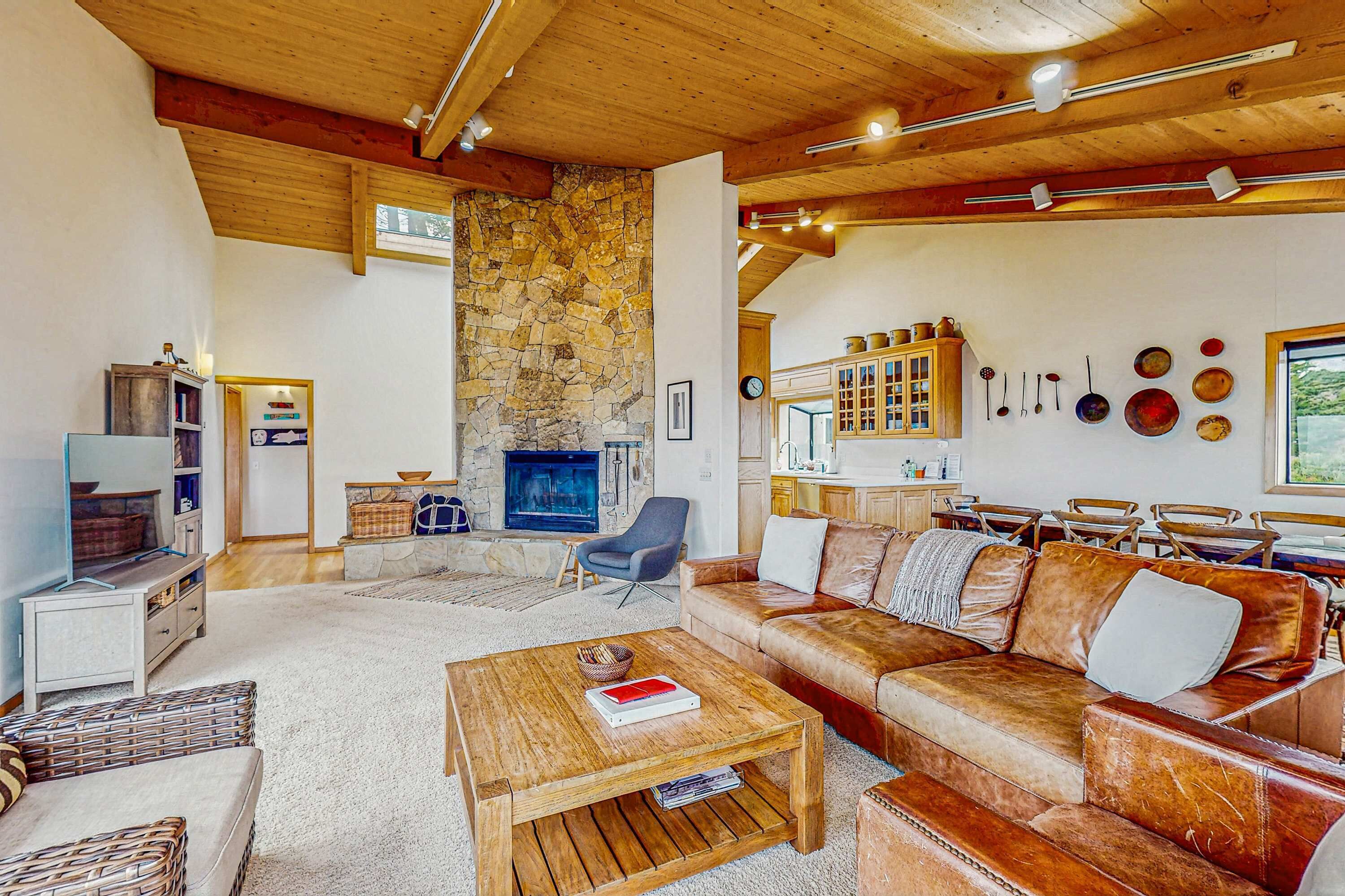 Un salon chaleureux avec une cheminée en pierre, des poutres apparentes en bois au plafond, un canapé en cuir et un coin repas avec des chaises en bois.
