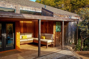 Eine Holzterrasse mit einer Sitzecke, Kissen, einer Schiebetür und einem kleinen Garten.