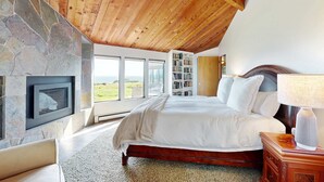 A bedroom with a stone fireplace, a large bed, a bookshelf, and a view of the outdoors. - Sea Ranch 4BR oceanfront home with private hot tub near Shell Beach (Sea Ranch)