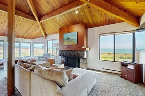 A spacious living room with a stone fireplace, large windows, and a high wooden ceiling. - Oceanfront home with private hot tub near Shell Beach (Sea Ranch)