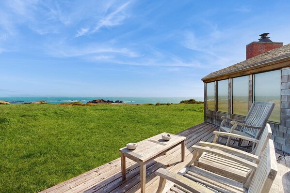 A wooden deck with a table and chairs, overlooking a grassy field and the ocean. - Oceanfront home with private hot tub near Shell Beach (Sea Ranch)