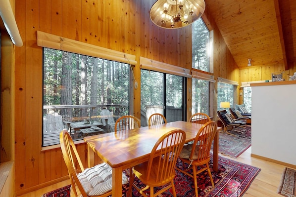 Un intérieur en bois avec une table à manger, des chaises et de grandes fenêtres donnant sur une forêt.