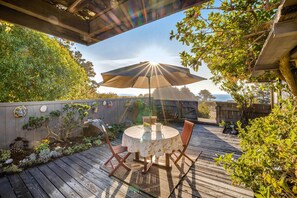 A wooden deck with a round table and chairs, an umbrella, and a garden with various plants and a wooden fence. - Esherick-designed home with private hot tub near Black Point, dogs welcome (Sea Ranch)