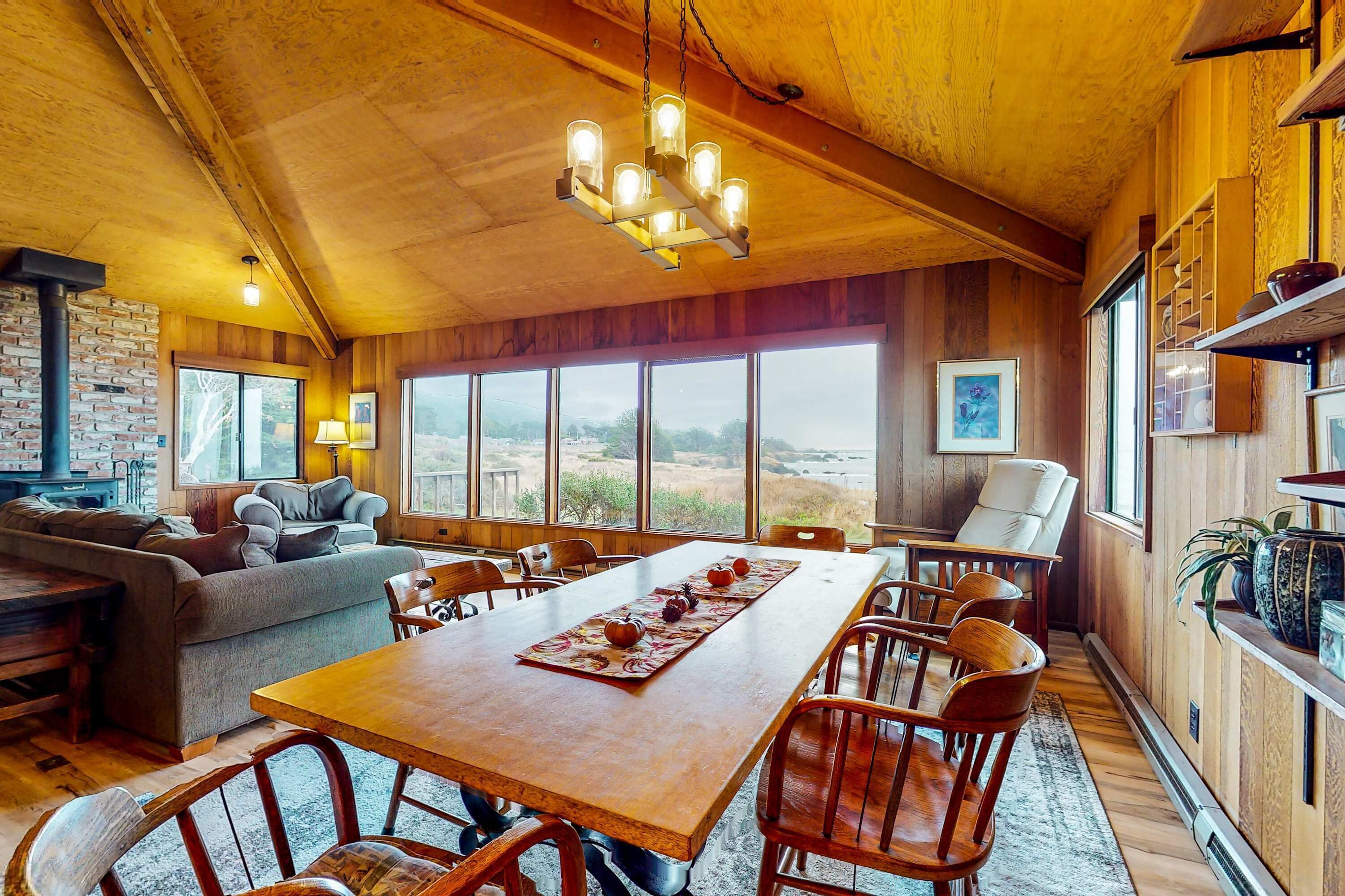 Un vaste espace dédié à la salle à manger, avec une longue table et des chaises en bois, une cheminée et de grandes fenêtres offrant une vue sur l’extérieur.