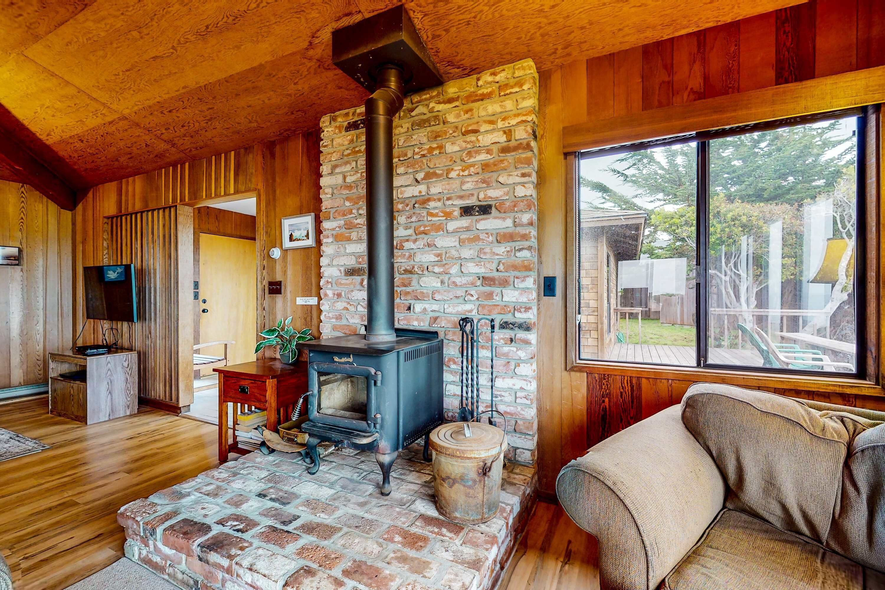 A cozy room with a brick fireplace, wooden walls, a sofa, and a large window.