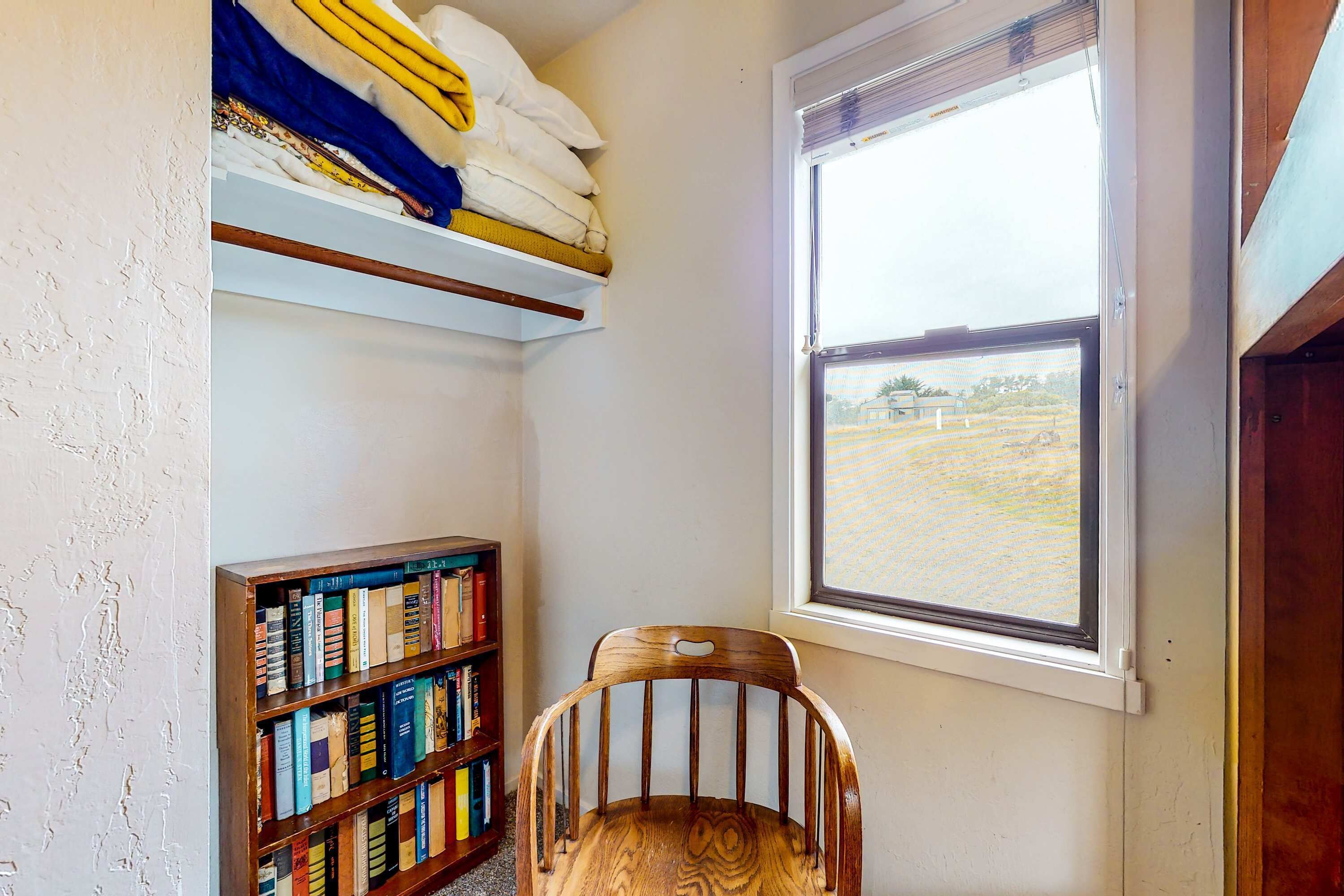 A room with a wooden chair, a bookshelf, a window with a view of a field, and a shelf with folded blankets.