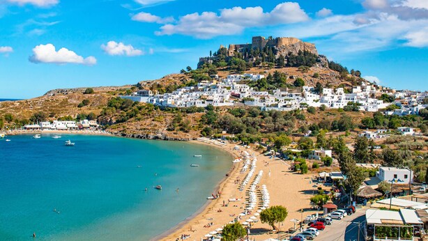 Une ville côtière avec un château perché sur une colline, une plage parsemée de parasols et des bateaux sur l’eau.