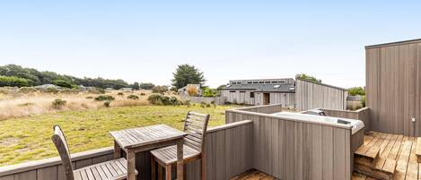 A deck with a wooden table and chairs, a hot tub, and a view of a grassy field and buildings in the distance.