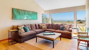 A modern living room with a sectional sofa, a coffee table, and a large window offering a view of the outdoors. - Sea Ranch 2BR modern ocean-view home on golf course with hot tub (Sea Ranch)