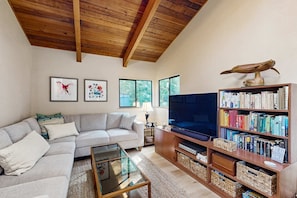 A living room with a sofa, a glass coffee table, a television, a bookshelf, and a wooden ceiling. - Meadow home with private hot tub near Black Point Beach (Sea Ranch)