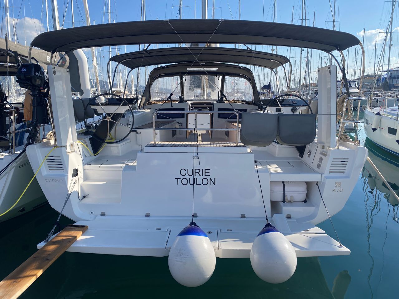 Ein Boot namens 'CURIE TOULON' liegt in einer Marina festgemacht, im Hintergrund sind weitere Boote zu sehen.
