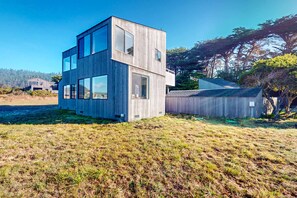 A modern wooden building with multiple floors and large windows, situated in a grassy area with trees in the background. - Dog-Friendly Sea Ranch Home Near Playground & Beach (Sea Ranch)