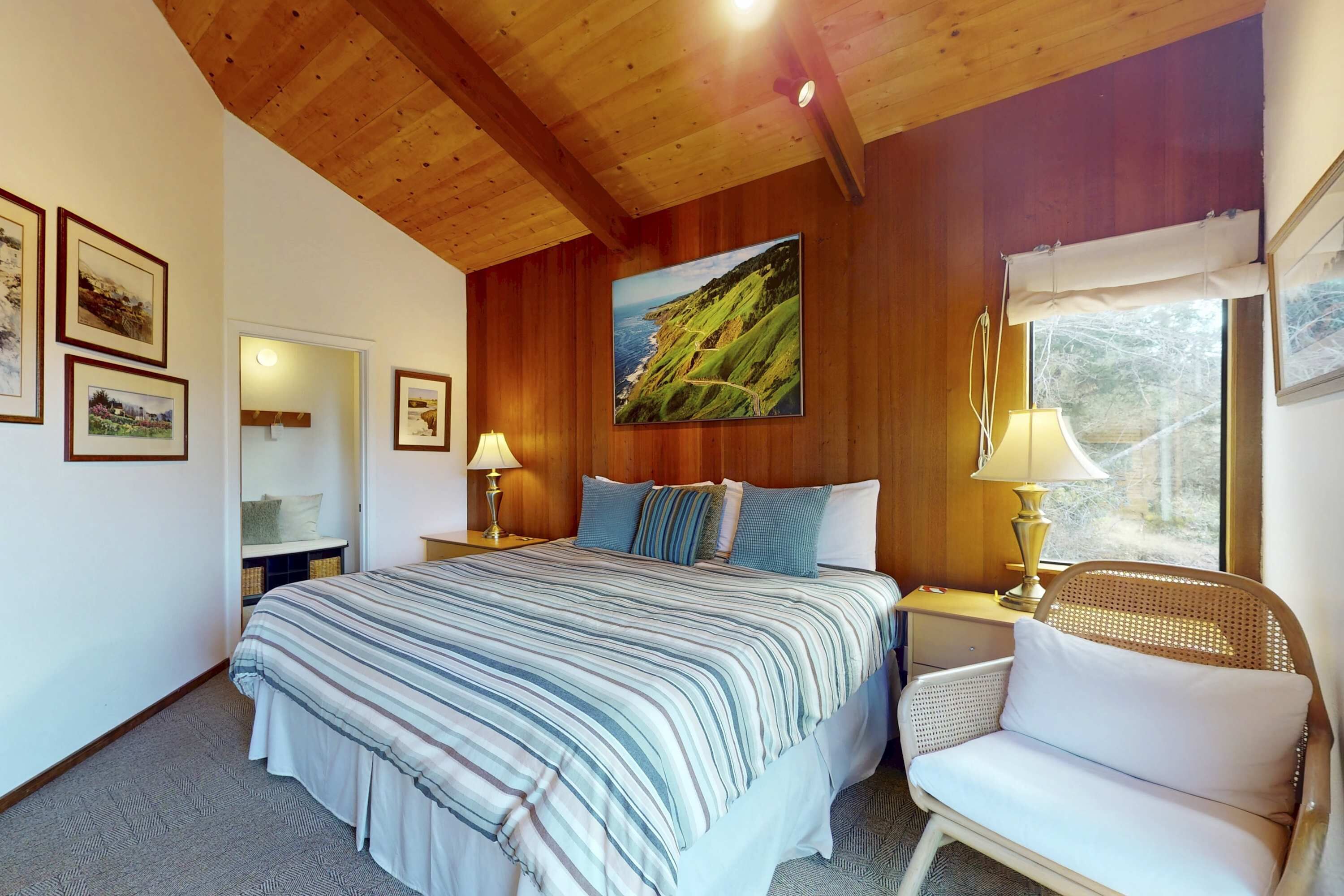 Quarto com cama, cadeira, mesa de cabeceira, luminária, janela e um quadro na parede.