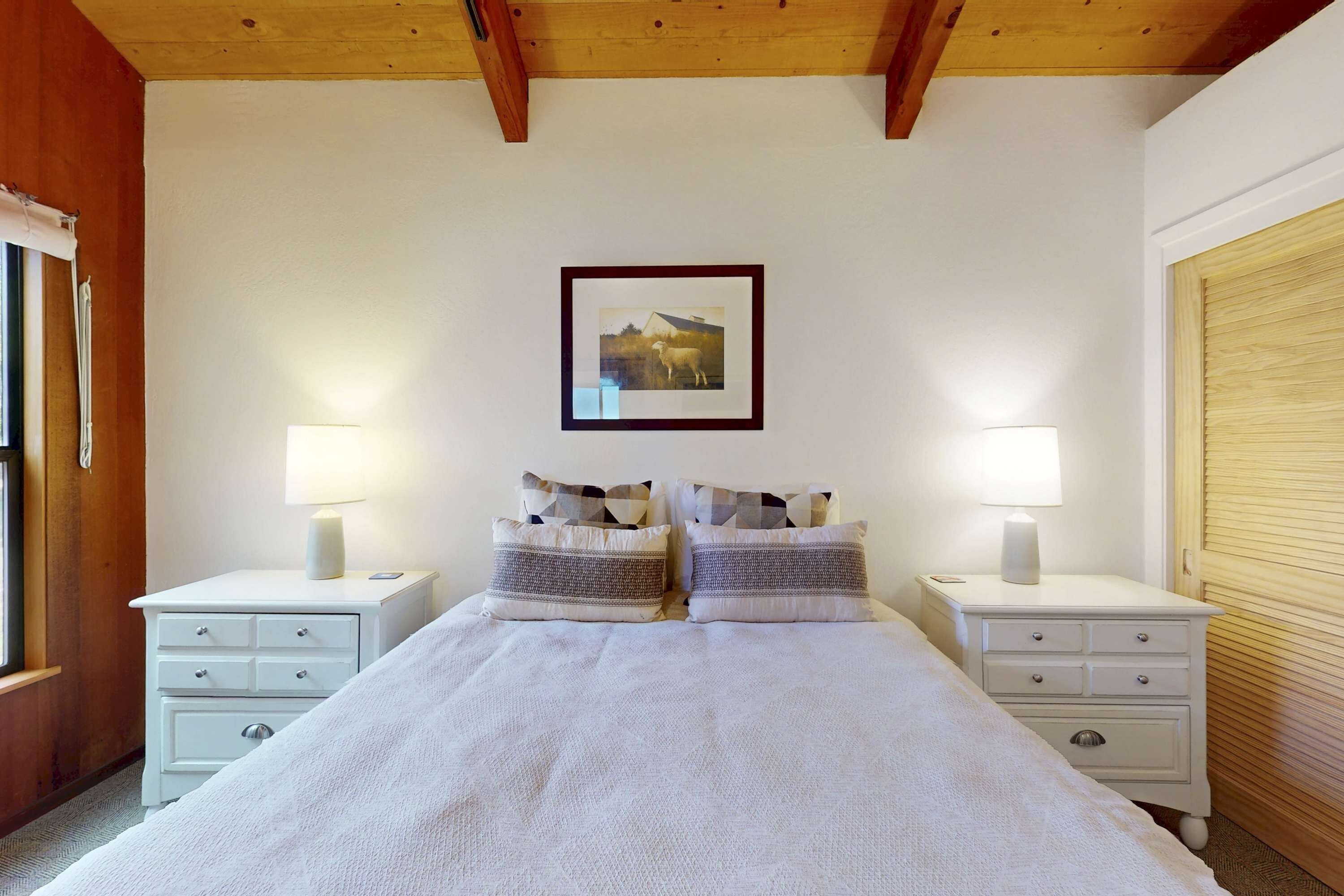 A bedroom with a large bed, two bedside tables with lamps, a framed picture on the wall, and a wooden ceiling.