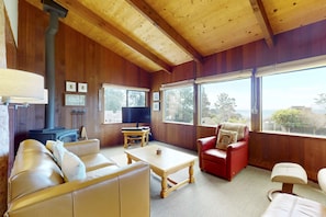 A cozy room with a sofa, a red chair, a wooden coffee table, a television, and a large window with a view. - Classic Sea Ranch Home With Updated Amenities. Hot Tub. 1 Dog Allowed (Sea Ranch)