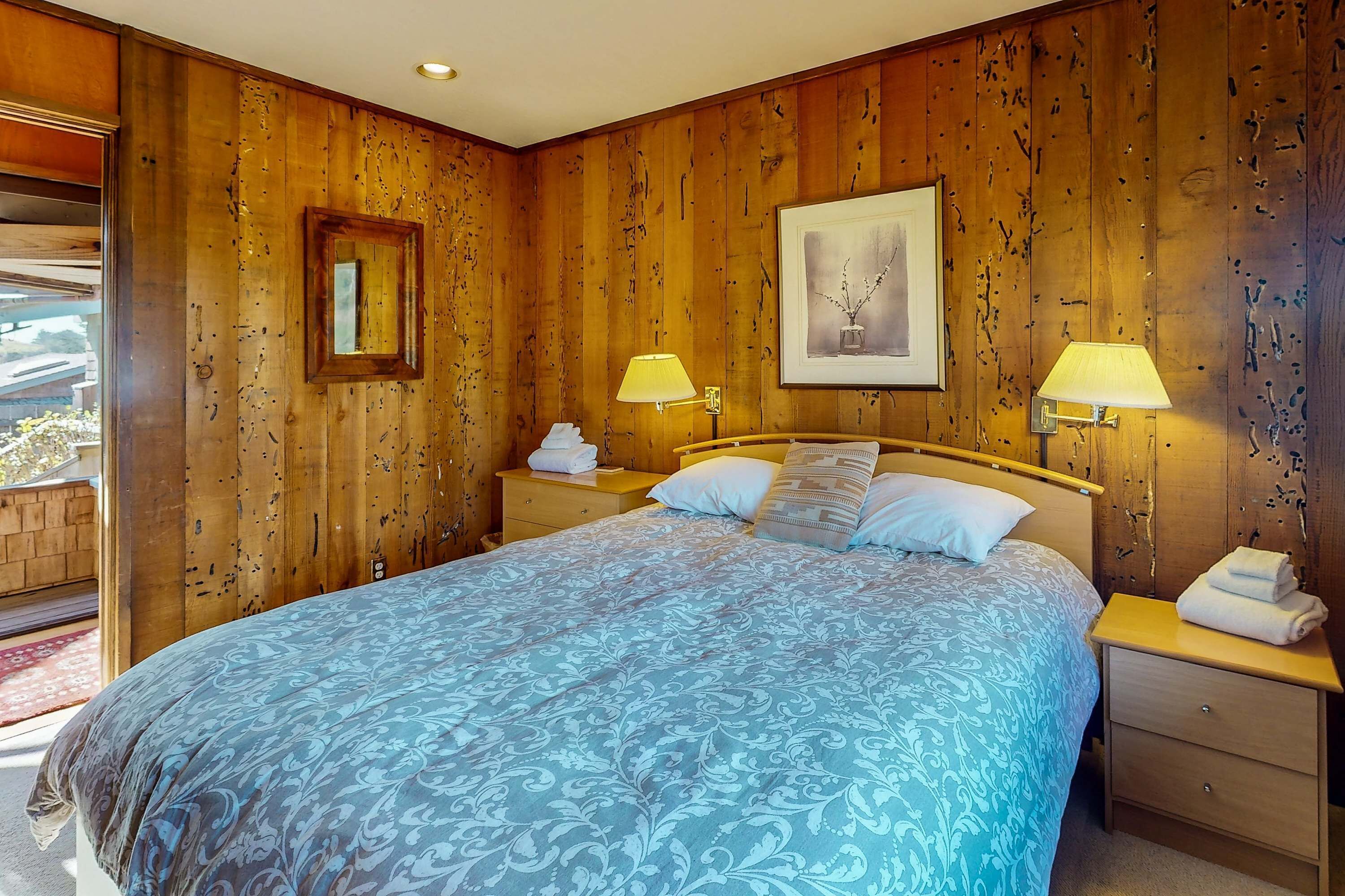Quarto com painéis de madeira, uma cama, duas luminárias de cabeceira, um quadro emoldurado e uma mesa de cabeceira com uma toalha.