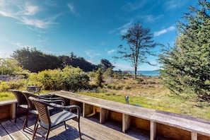An outdoor deck with a long wooden table and wicker chairs, overlooking a natural landscape with trees and a view of the ocean. - Ocean-view home with hot tub near Black Point (Sea Ranch)